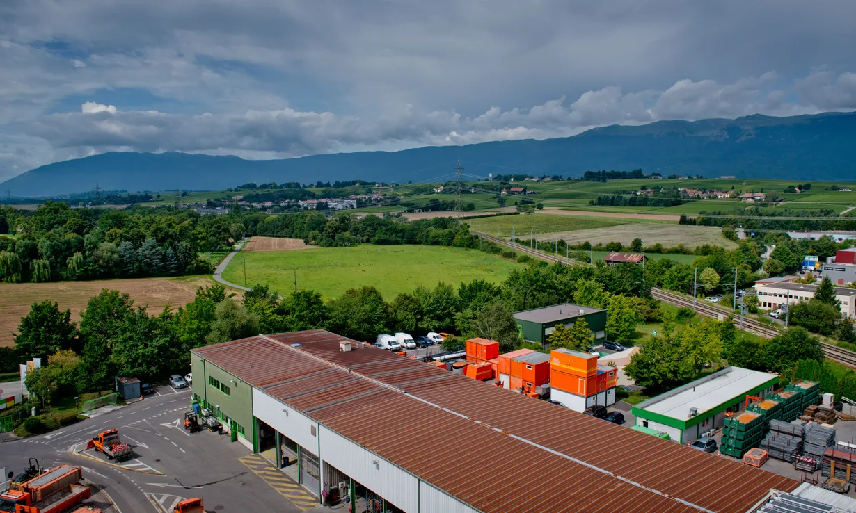 Vue aérienne d'un bâtiment industriel entouré de champs, avec le Jura à l'horizon